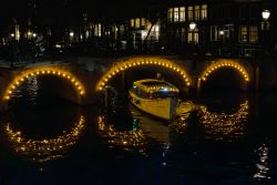 Amsterdam und Umgebung April 2025 Ein beleuchtetes Boot fährt unter einer funkelnden Brücke in Amsterdam durch Amsterdam und Umgebung April 2025 Abendliche Kanalansicht in Amsterdam mit beleuchtetem Boot und Brücke