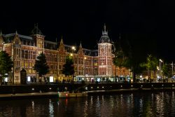 Amsterdam und Umgebung April 2025 Nachtaufnahme des eindrucksvollen Bahnhofs Amsterdam Centraal und seiner Spiegelung im Wasser. Amsterdam und Umgebung April 2025 Beleuchteter Bahnhof Amsterdam Centraal bei Nacht mit reflektierendem Wasser