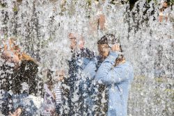 Amsterdam und Umgebung April 2025 Eine Gruppe von Menschen steht hinter einem spritzenden Springbrunnen Amsterdam und Umgebung April 2025 Menschen genießen Springbrunnen, durch spritzendes Wasser verdeckt