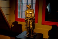 Amsterdam und Umgebung April 2025 Eine goldene Statue steht in einem beleuchteten Raum vor einem Fenster Amsterdam und Umgebung April 2025 Goldene Statue eines Mannes in traditioneller Kleidung vor einem Fenster