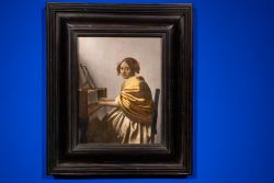 Amsterdam und Umgebung April 2025 Die Komposition zeigt eine Frau, die am Cembalo sitzt, umgeben von einem prächtigen Rahmen Amsterdam und Umgebung April 2025 Gemälde einer Frau am Cembalo, eingerahmt und an blauer Wand