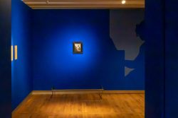 Amsterdam und Umgebung April 2025 Ein Kunstwerk zieht die Aufmerksamkeit in einem intensiv blau gestalteten Museumsraum auf sich. Amsterdam und Umgebung April 2025 Blau beleuchtetes Kunstmuseum mit kleinem gerahmtem Gemälde und Schatten eines Betrachters