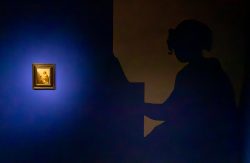 Amsterdam und Umgebung April 2025 Ein Gemälde leuchtet in einem dunklen Raum neben der Silhouette einer Klavierspielerin Amsterdam und Umgebung April 2025 Silhouette einer Frau am Klavier, beleuchtetes Gemälde an blauer Wand