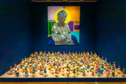 Amsterdam und Umgebung April 2025 Eine Kunstinstallation zeigt zahlreiche bunte Figuren unter einem leuchtenden Porträt an einer blauen Wand. Amsterdam und Umgebung April 2025 Moderne Kunstinstallation mit bunten Figuren vor einem farbenfrohen Porträt