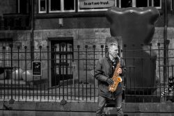 Fotografie von Aloys Peter Trenz Straßenmusiker spielt Saxophon in einer urbanen Umgebung mit moderner Kunst Ausgewählte Fotomotive von Aloys Peter Trenz