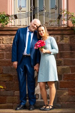 Hochzeitsfotografie Glückliches Paar in eleganter Kleidung vor Backsteinmauer, sie hält einen Blumenstrauß