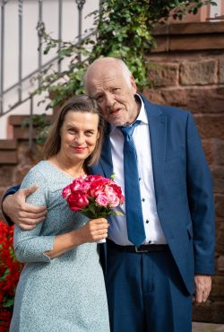 Hochzeitsfotografie Älteres Paar in eleganter Kleidung vor steinernem Gebäude mit Blumenstrauß
