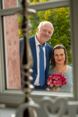 Hochzeitsfotografie Älteres Paar lächelt durch ein Fenster, Frau hält roten Blumenstrauß