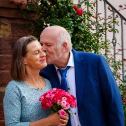 Hochzeitsfotografie Älteres Paar mit Blumen vor rosengewachsenem Hintergrund in einer liebevollen Pose