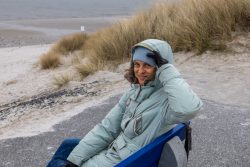 Insel Sylt Februar 2025 Eine Frau genießt die kalte Brise am Strand vor den Dünen Insel Sylt Februar 2025 Frau in blauer Winterjacke sitzt am Strand mit Dünen im Hintergrund