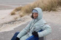 Insel Sylt Februar 2025 Eine Frau genießt eine Pause auf einer Bank an einem winterlichen Strand mit Dünen im Hintergrund Insel Sylt Februar 2025 Frau in Winterjacke sitzt auf einer Bank am Strand mit Dünen