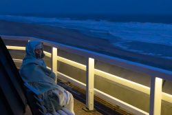 Insel Sylt Februar 2025 Eine Frau sitzt abends eingehüllt in eine Decke auf einer Bank an der Küste. Insel Sylt Februar 2025 Frau in Decke gehüllt auf nächtlicher Strandpromenade mit Blick auf das stürmische Meer