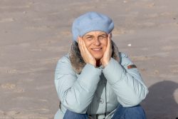 Insel Sylt Februar 2025 Eine ältere Frau genießt einen sonnigen Tag am Strand in Winterkleidung Insel Sylt Februar 2025 Ältere Frau im blauen Mantel und Barett am Strand sitzend