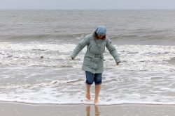 Insel Sylt Februar 2025 Eine Frau genießt einen Spaziergang am Strand, trotz der kühlen Brise Insel Sylt Februar 2025 Frau im blauen Mantel spaziert barfuß am Strand entlang