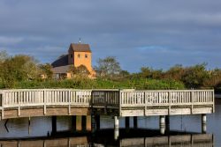 Insel Sylt und Hallig Hooge Oktober 2025 Ein Holzsteg führt über einen ruhigen See mit einer Kirche im Hintergrund Insel Sylt und Hallig Hooge Oktober 2025 Holzsteg mit Kirche im Hintergrund an einem ruhigen See