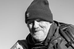 Insel Sylt und Hallig Hooge Oktober 2025 Ein älterer Mann in warmer Kleidung genießt die Natur Insel Sylt und Hallig Hooge Oktober 2025 Schwarzweißfoto eines lächelnden älteren Mannes mit Mütze und Rucksack