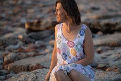 Portraitfotografie Fotoshooting Strand Helgoland August 2024 Frau in buntem Sommerkleid sitzt auf felsigem Strand bei Sonnenuntergang