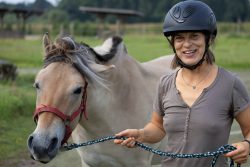 Reitstunde Fotografie Aloys Peter Trenz Frau mit Reithelm steht neben einem Pferd auf einer Wiese