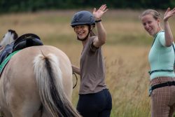 Reitstunde Fotografie Aloys Peter Trenz Zwei Frauen winken auf einer Pferdekoppel mit einem Pferd