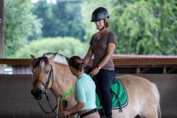 Reitstunde Fotografie Aloys Peter Trenz Frau lernt Reiten in Reithalle mit braunem Pferd