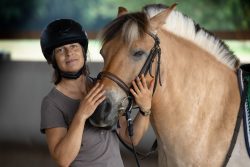 Reitstunde Fotografie Aloys Peter Trenz Frau mit Helm posiert neben einem Fjordpferd in einem Reitstall