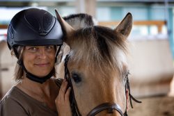 Reitstunde Fotografie Aloys Peter Trenz Frau mit Helm neben einem Pferd im Reitstall