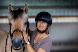 Reitstunde Fotografie Aloys Peter Trenz Frau mit Helm lächelt neben einem Pferd in einer Halle